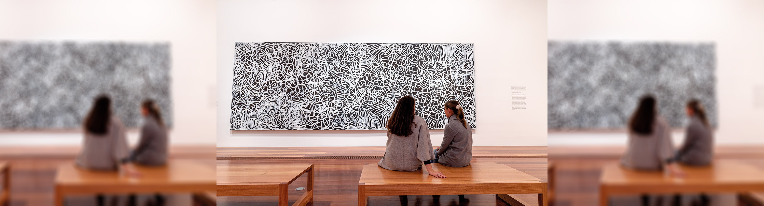 Two women sitting on a bench in The Ian Potter Centre: NGV Australia looking at a painting