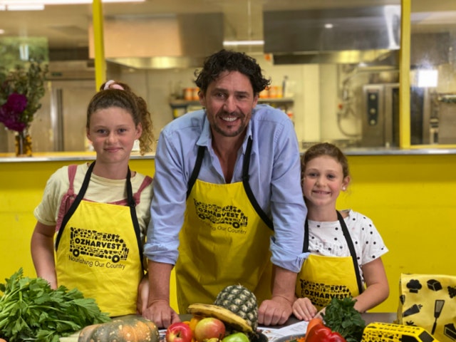 Chef Colin Fassnidge is standing in between his daughters who are all wearing yellow aprons and are smiling