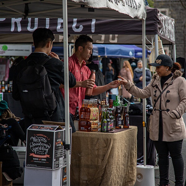 Two people are standing behind a counter under a marquee for a cider ccompany and are handing a tasting to a woman wearing a duffel jacket and cap