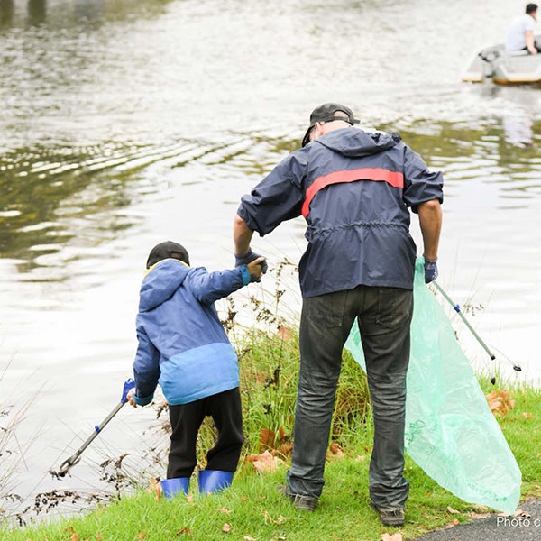 A man and a child have their backs to the camera and are wearing rain coats, both are holding rubbish grabbers and the man is holding the child's hand while the child is using the grabber to reach into the Yarra River