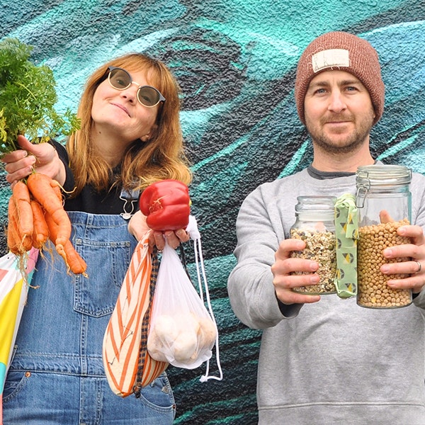 A woman in denim overalls is holding a bunch of carrots in one hand and a capsicum and plastic bags in the other, on her left is a man wearing a beige beanie and grey jumper holding a jar of lentils in each hand