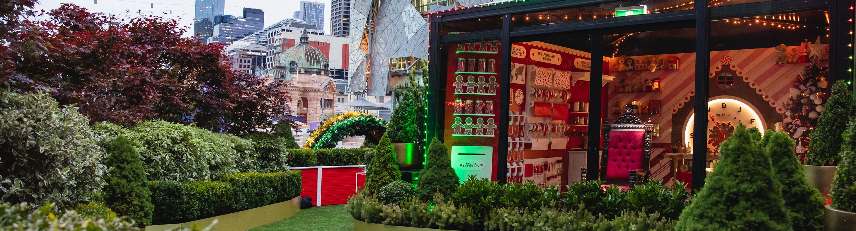 A photo of Santa's workshop with a bright red chair in the window and the workshop is surrounded by greenery in Fed Square