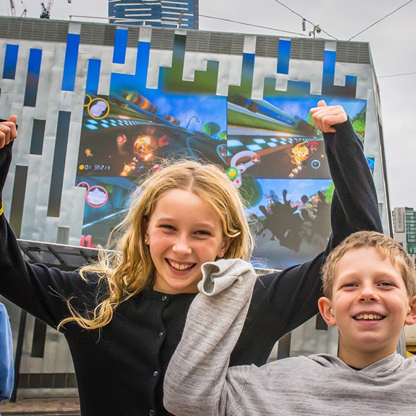 Two kids cheering in front of the digital facade while Mario Kart is being played behind them