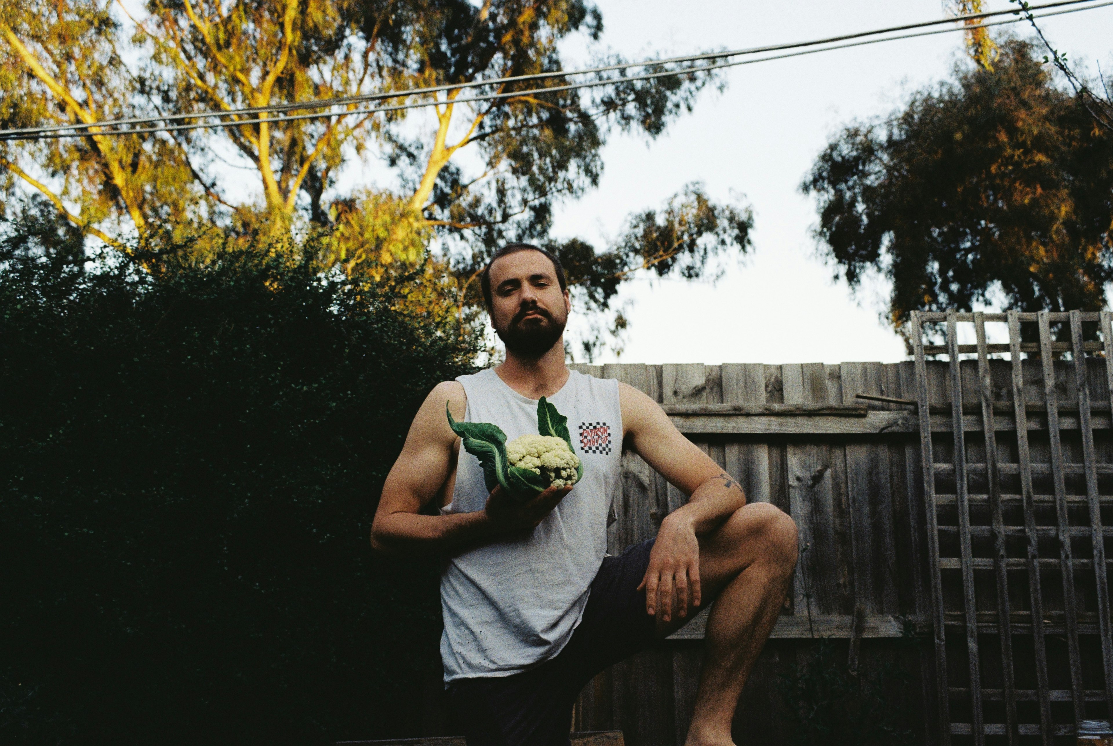 Ryan Prehn stands in a garden, in front of a fence. He wears a white sleeveless tee and hold a cauliflower as he looks at the camera. His arm is resting on his raised knee.