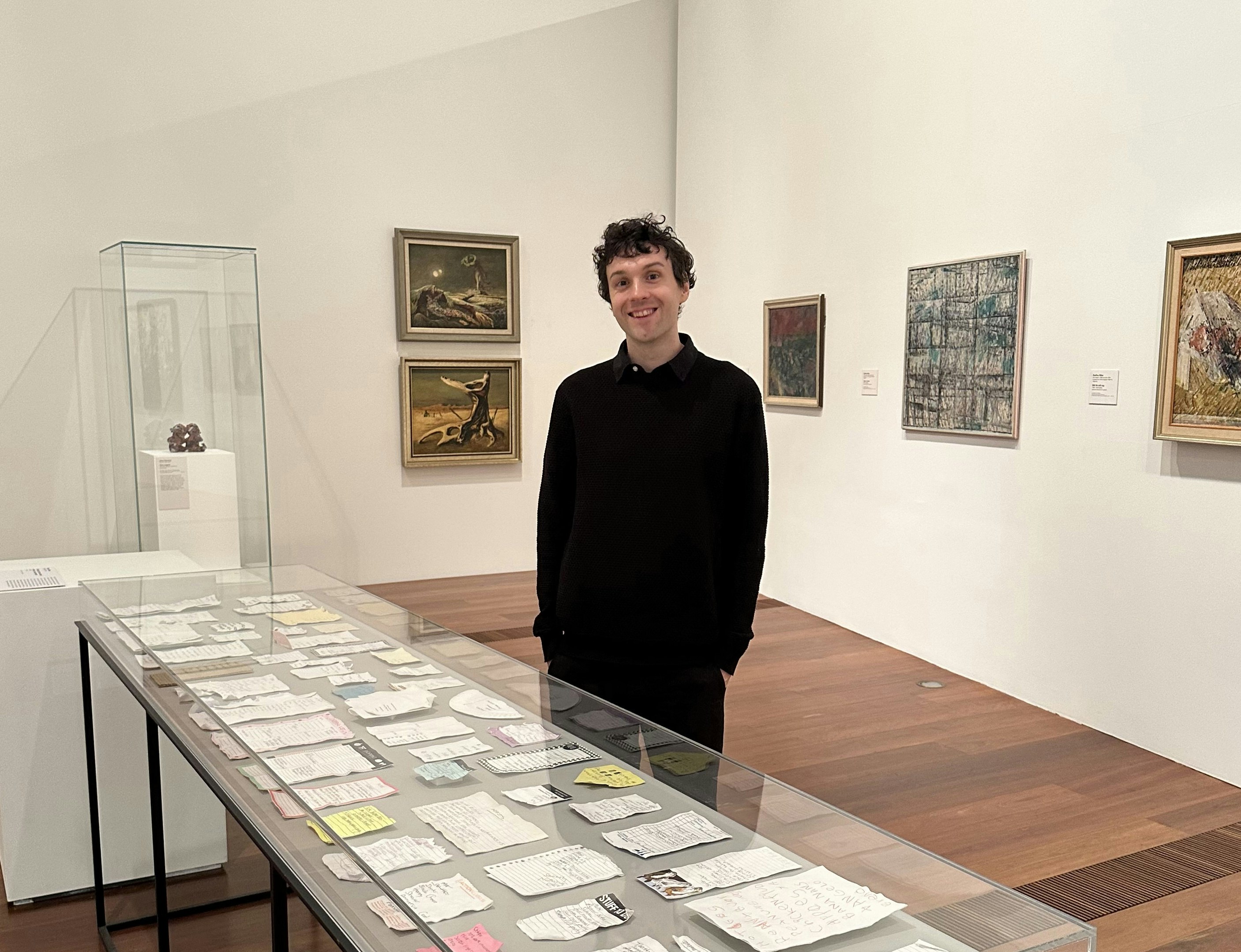 Kenny Pittock, dressed in black, stands behind a glass display cabinet with a series of ceramic white lists. There is artwork hung on the exhibition walls behind him.