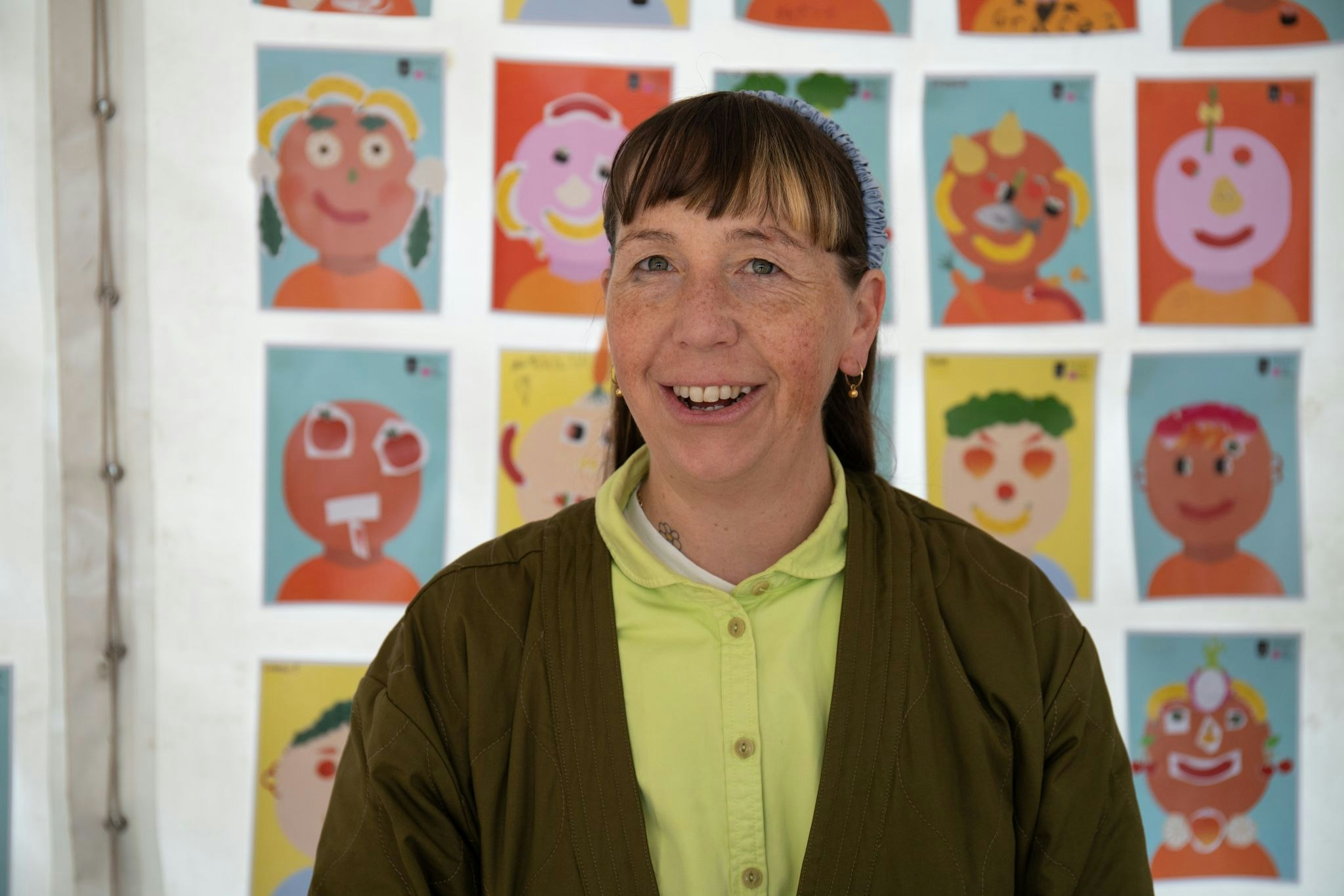 Artist Beci Orpin smiles at the camera. She wears a green jacket. Behind her hangs a series of food collage pictures by children.