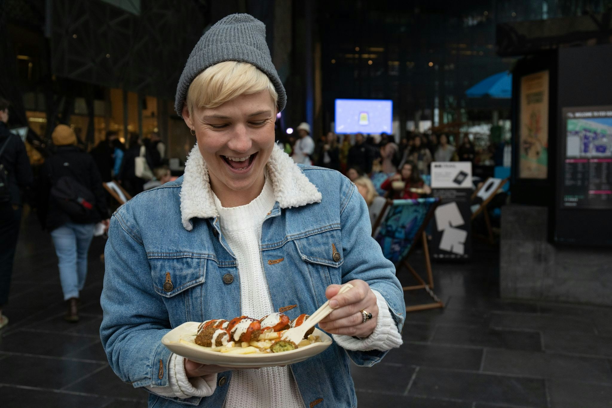 A woman wearing a grey beanie and a denim jacket eats a plate of falafel. A festival in the atrium at Fed Square is going on behind her, with stallholders selling things.