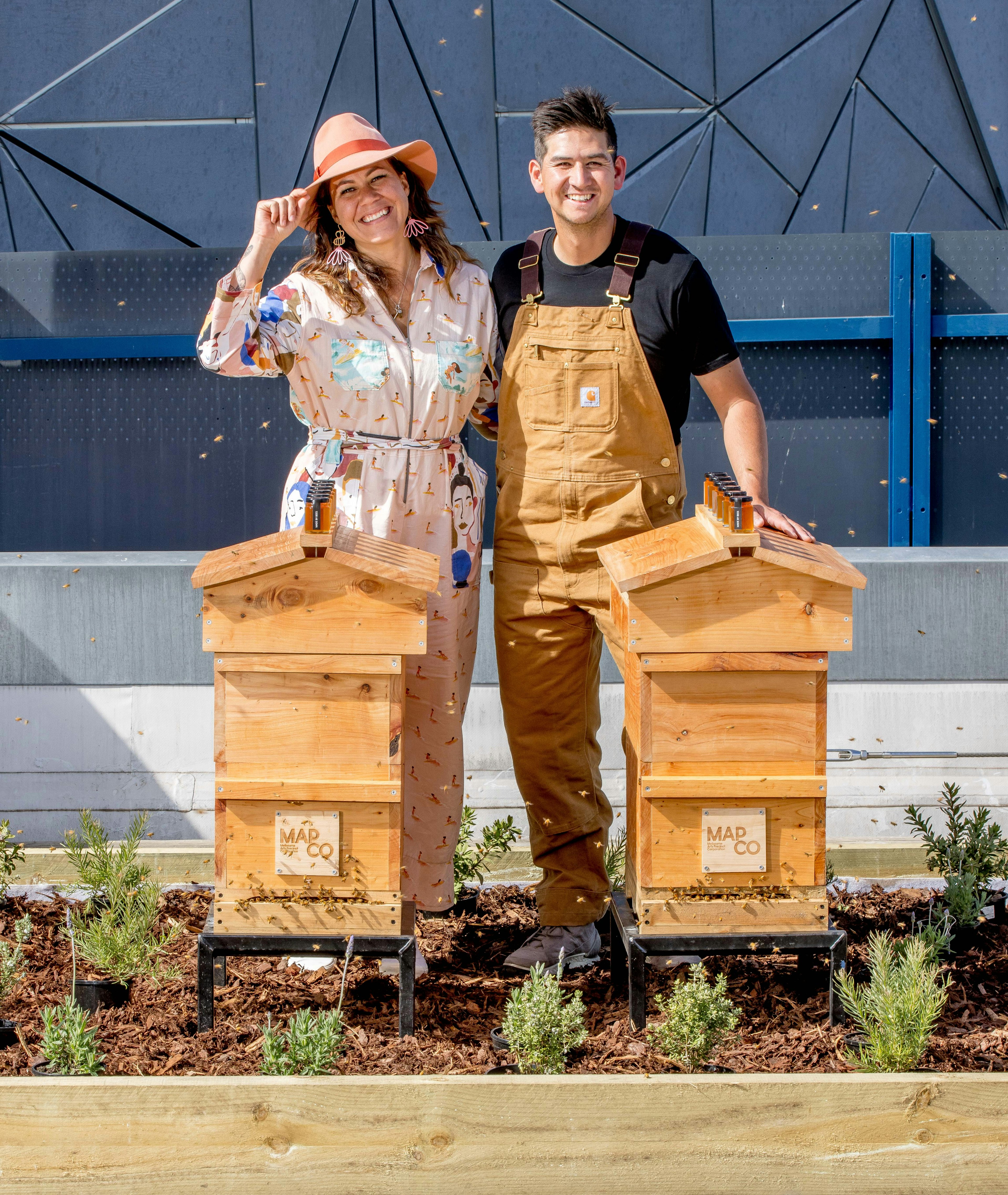 A man and woman stand in a garden bed, behind two timber beehives, smiling at the camera.