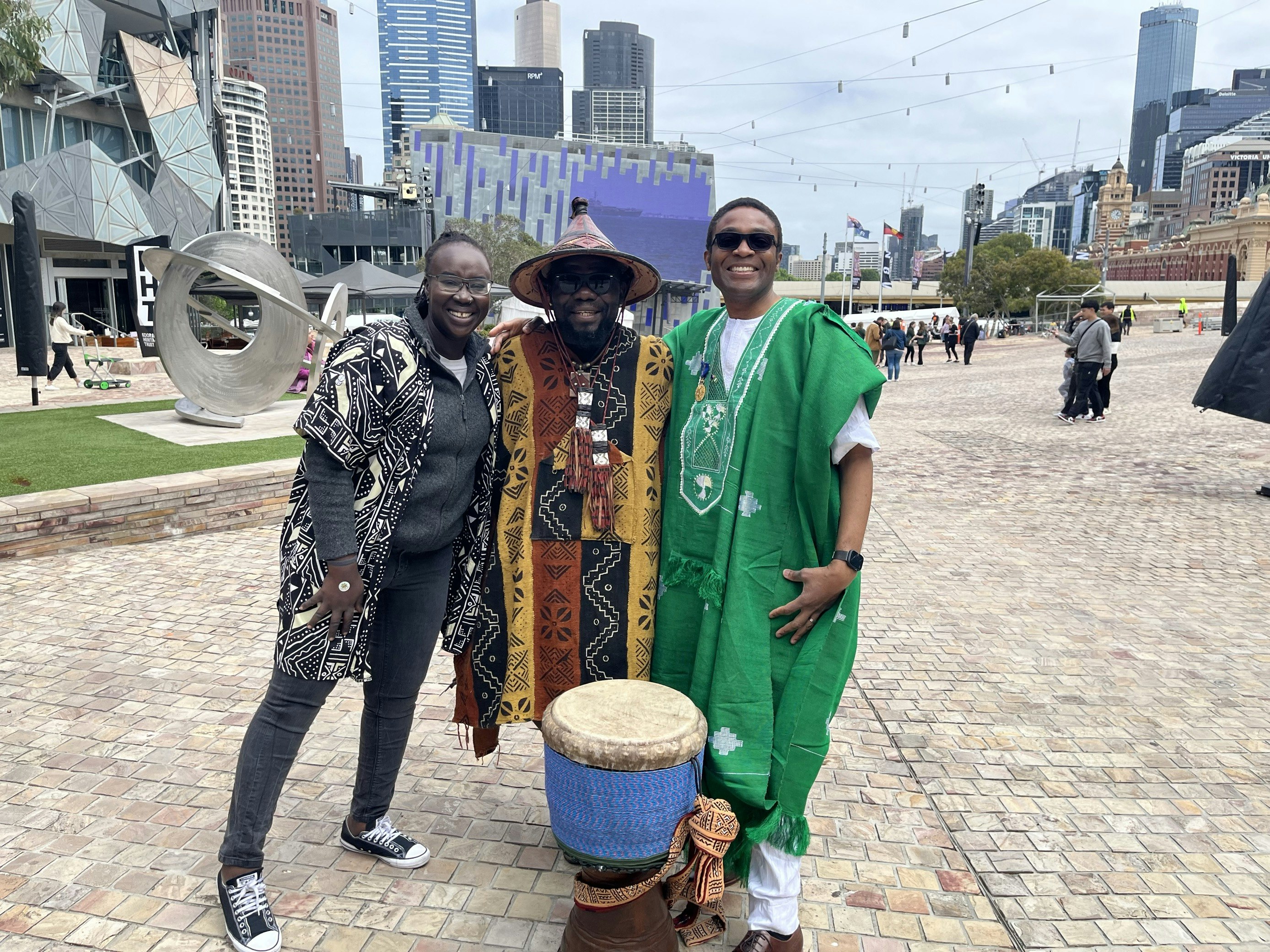 Poni Tungun, Kofi Kunkpe and Fred Alale AM at Fed Square. Poni is on the left, wearing a patterned black and white jacet, Kofi is in the centre, with an African drum in front of him, and wears a hat and traditional costume. Fred is on the right, and wears bright green robes and sunglasses. The Fed Square Big Screen is behind them.