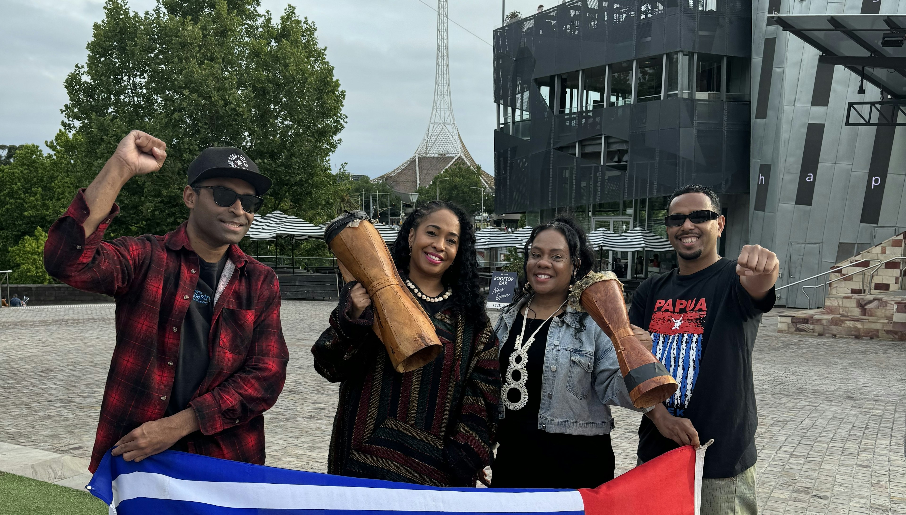 Ronny Kareni, The Black Sistaz and Sam Roem face the camera, smiling, the Arts Centre behind them. The Black Sistaz hold up tifa drums, and Ronny and Sam have their hand in the air, Ronny with his right fist raised. We see the top of a flag they are holding.
