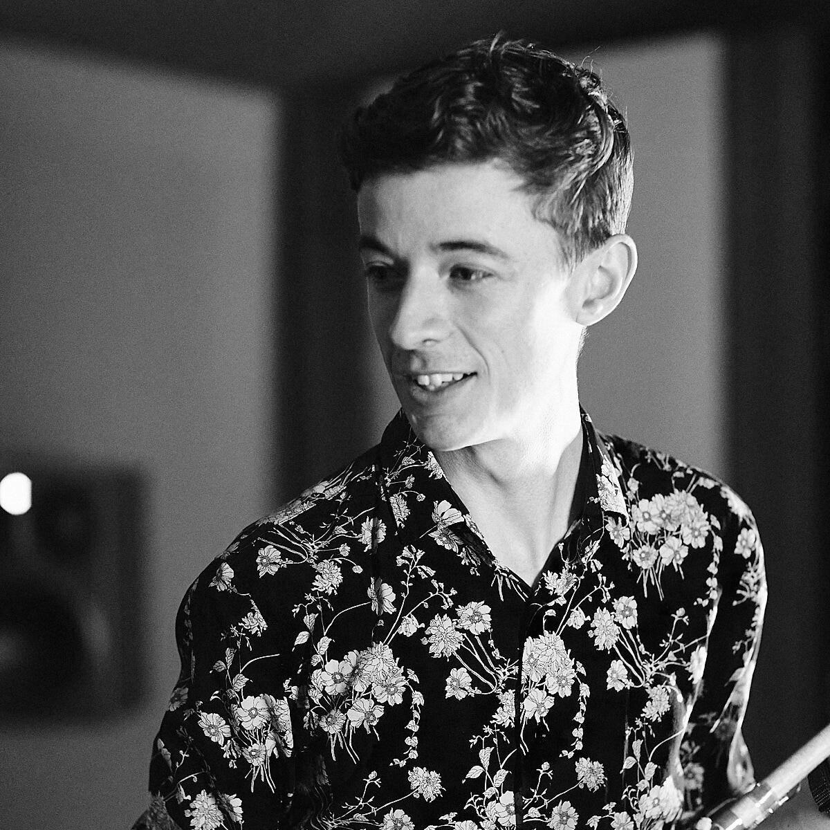 Composer Ciaran Frame wears a floral shirt, looking to the left of the camera. The image is black and white, and you can see part of his flute that he is holding