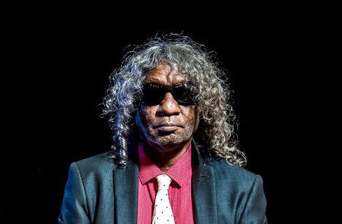 Uncle Bart wears black sunglasses, facing the camera. He has shoulder-length curly grey hair, wears a red shirt, white tie and charcoal jacket.