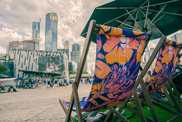 A photo of deck chairs under an umbrella with the screen at Fed Square in the background