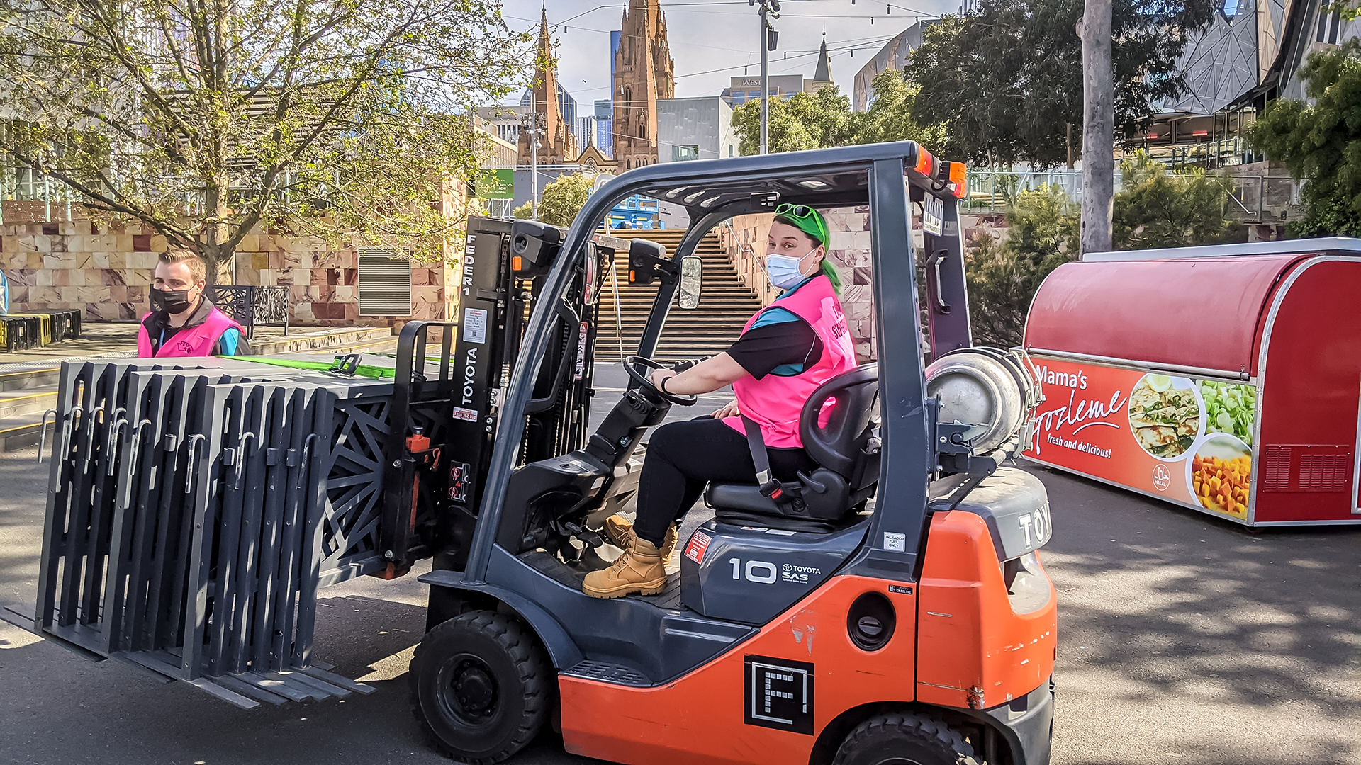 Ellora is sitting in a forklift that is facing left, looking at the camera in a fluoro pink vest