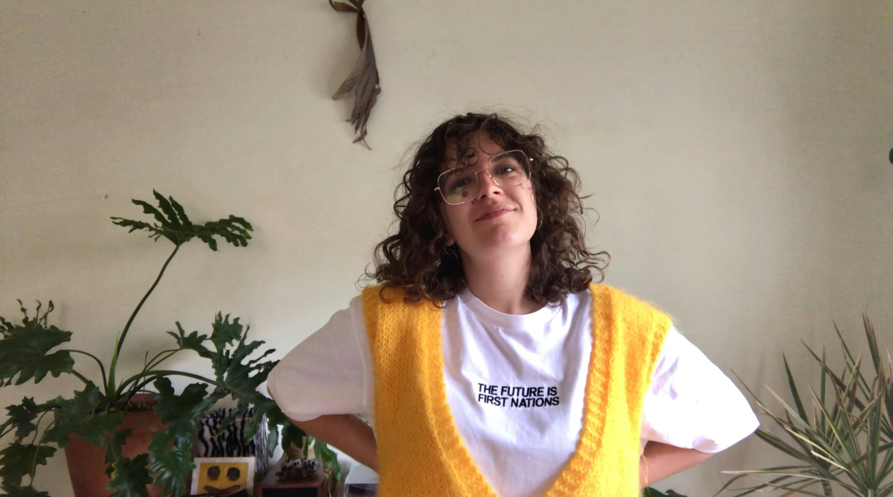 Pierra is standing in a room with their hands on their hips wearing a yellow vest and white T-shirt in front of some green plants