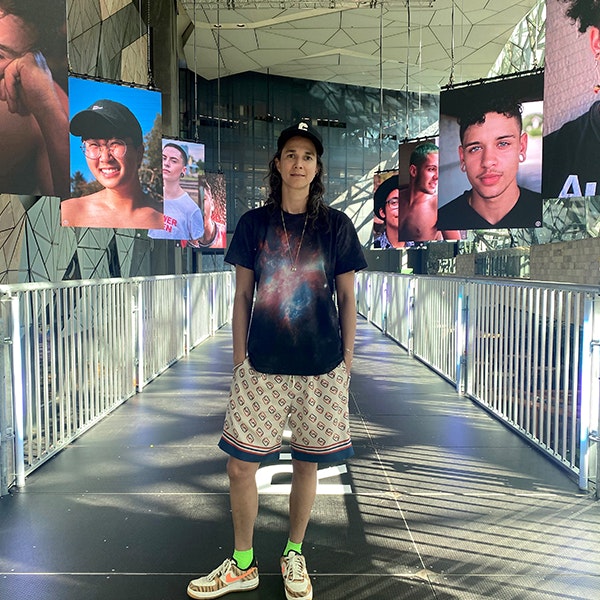 Soraya is standing on the bridge of the Reconstruct (the) Normative exhibition at Fed Square, they are wearing a cap, a black T shirt and check shorts