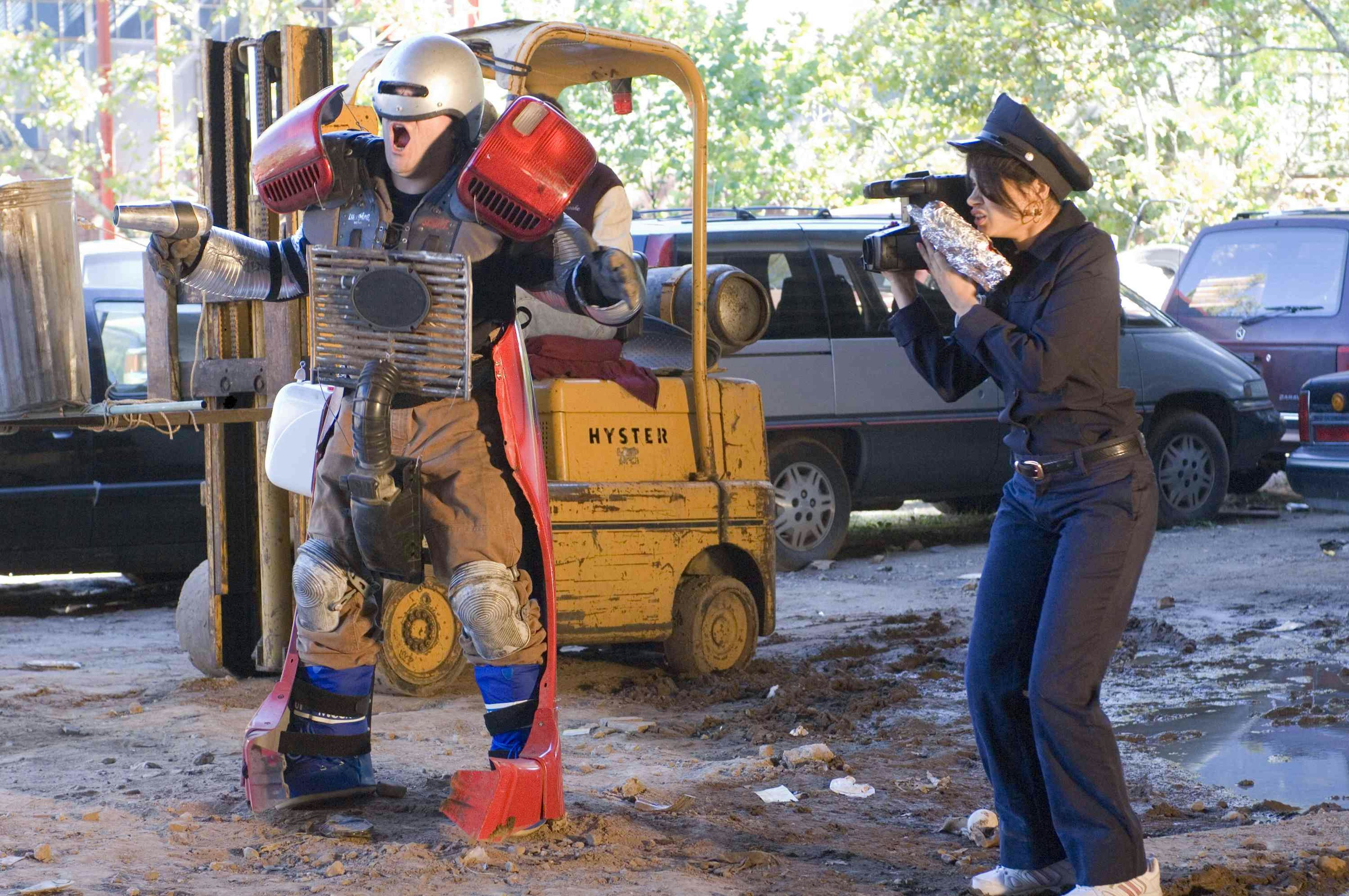 Jack is dressed as Robocop in front of a yellow forklift as a person dressed as police films him