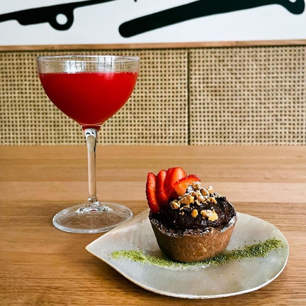 A photo of a red cocktail in a glass with a stem next to a small chocolate tart with pistachio dust on the plate and sliced strawberry on the chocolate