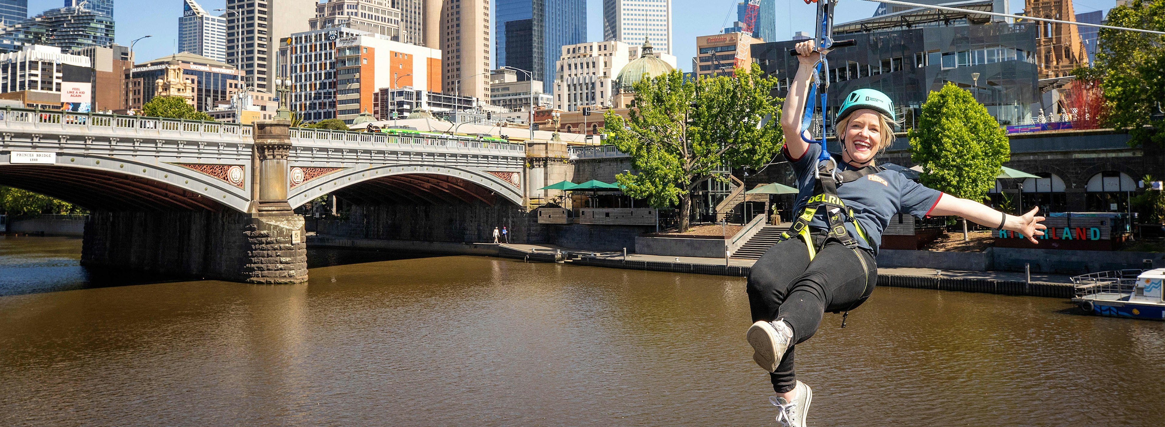 An image of a smiling person taking a zipline across the Yarra River near Princes Bridge