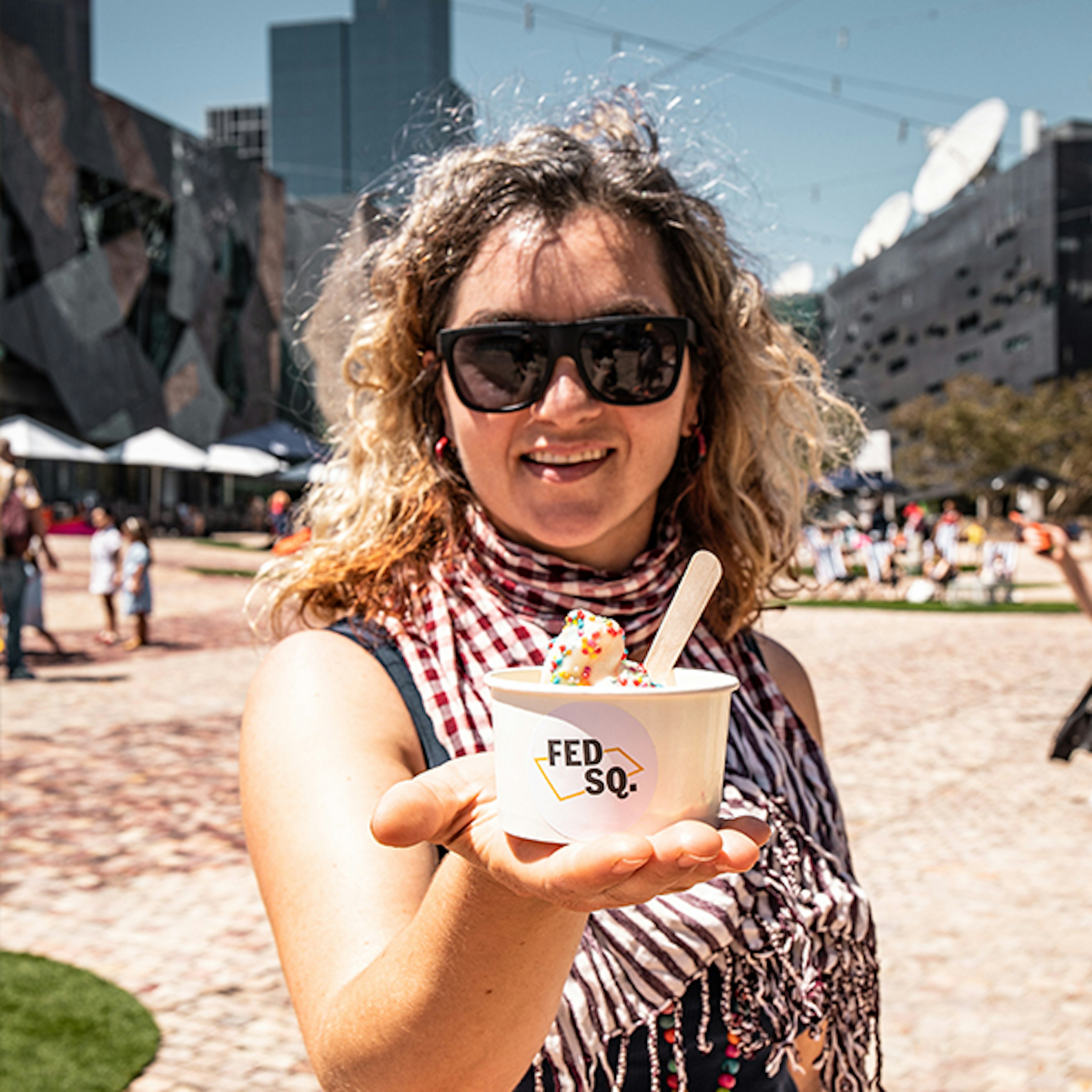 A woman wearing sunglass and holding a cup of ice cream while smiling and looking at the camera