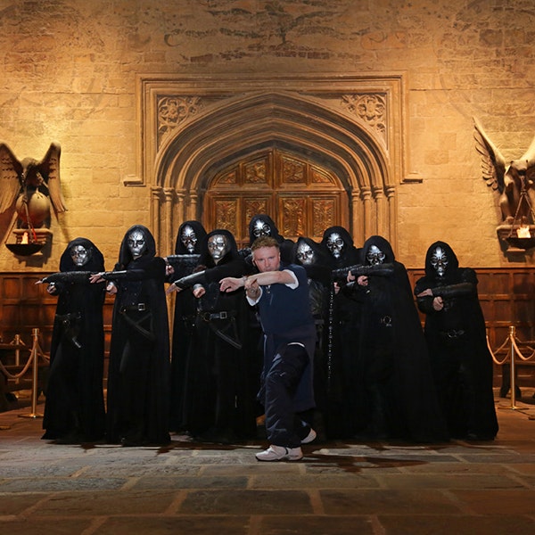 Paul Harris pointing his wand at the camera with a group of Death Eaters standing behind him in front of the doors to the great hall of Hogwarts