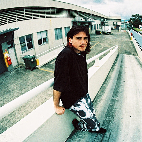Musician Chris Lanzon is standing behind some industrial buildings on a ramp looking at the camera wearing a black t-shirt