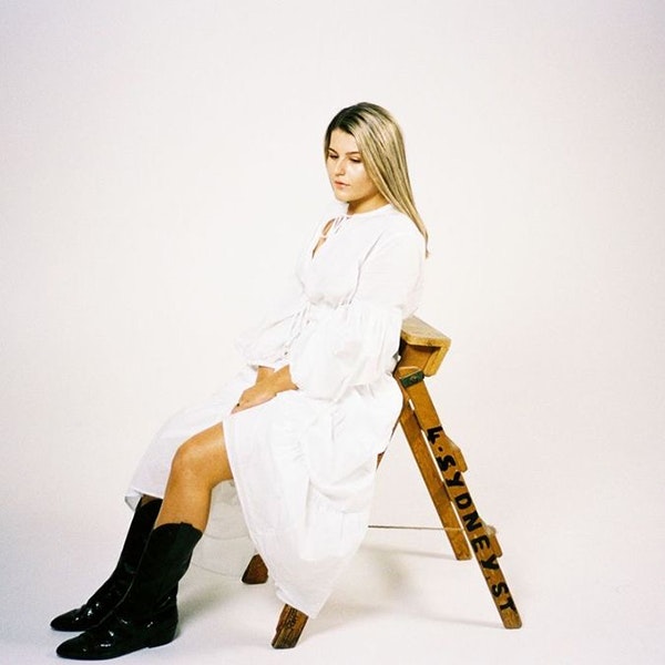 Musician Eliott is sitting profile view on a step ladder looking down wearing a white dress and black cowboy boots