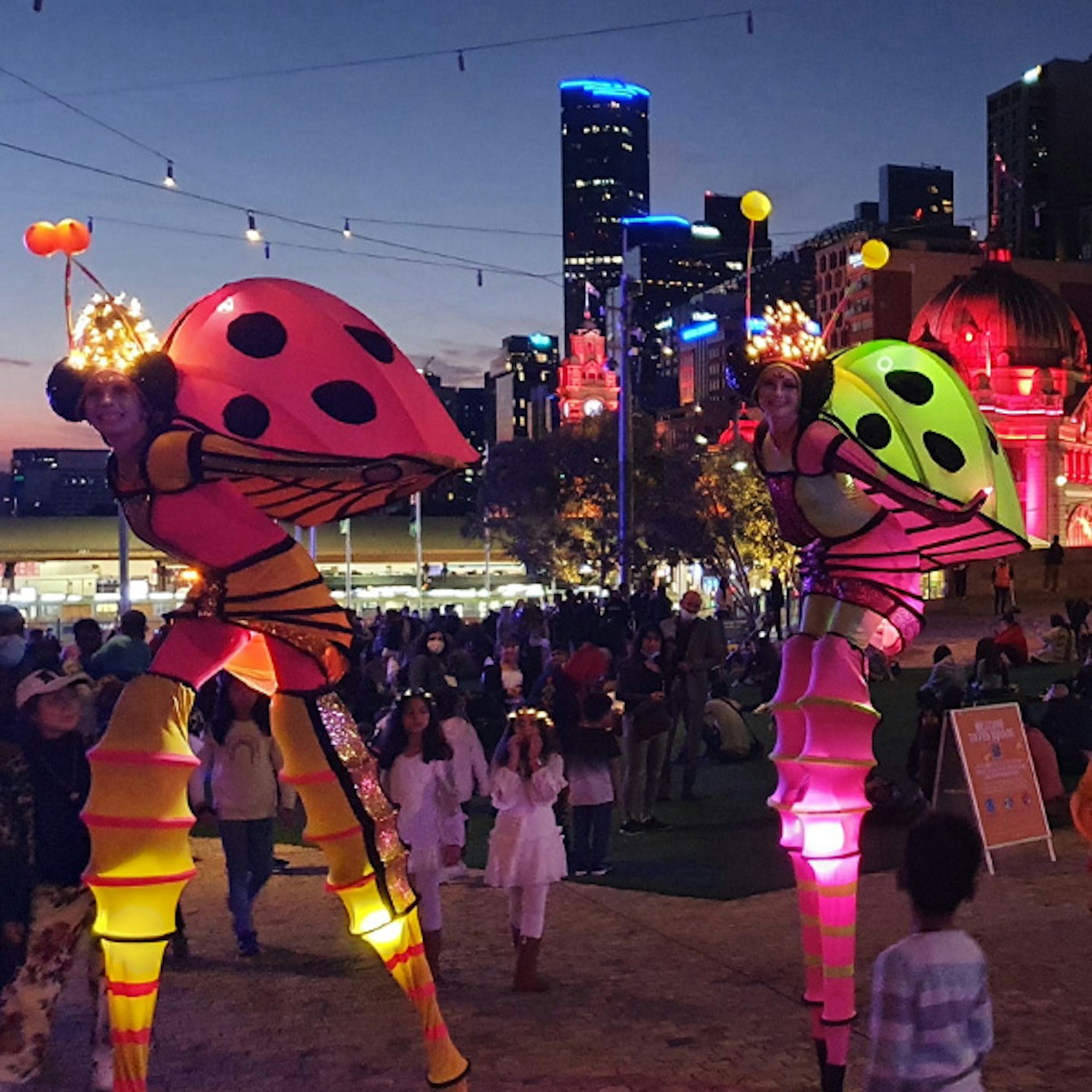 Two ladybug stilt walkers illuminated while walking through crowds at Fed Square