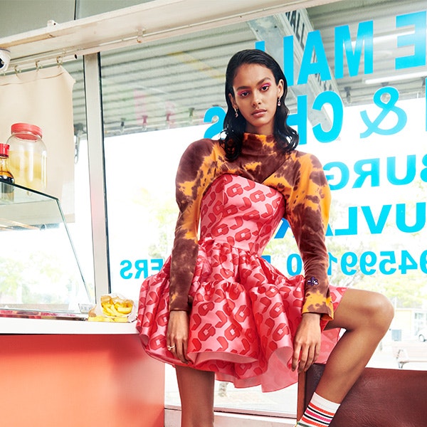 A model wearing a pink dress and tie dye long sleeve shirt, with her leg up on a chair in a fish and chip shop for Melbourne Fashion Festival