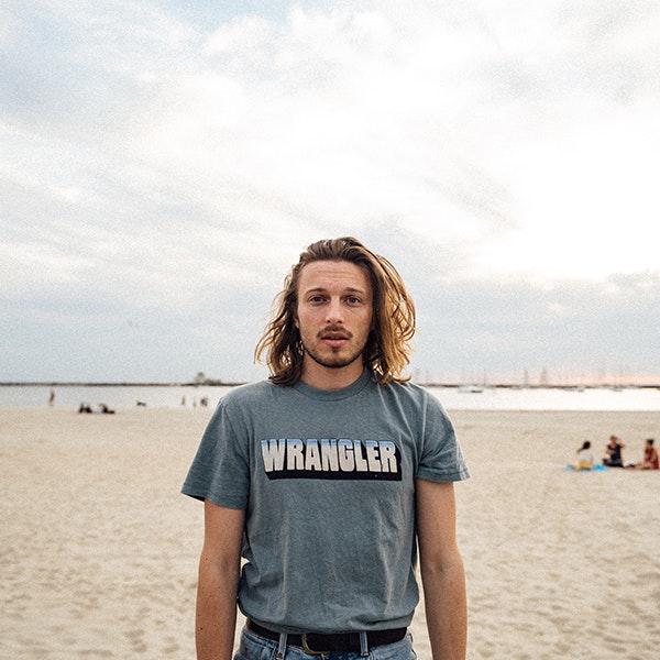 Singer Seb Szabo is looking at the camera while standing on a beach wearing a grey Rangler T Shirt