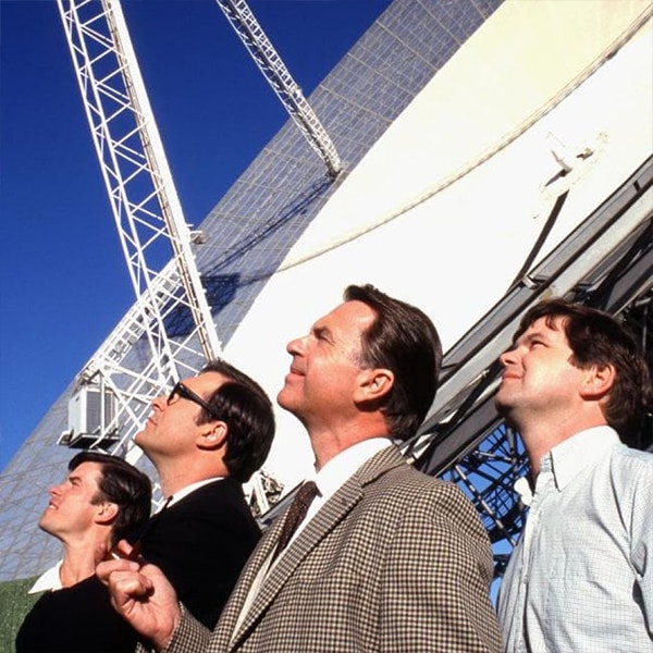 Sam Neill and his team in The Dish are standing outside looking at the sky with the large satellite dish behind them