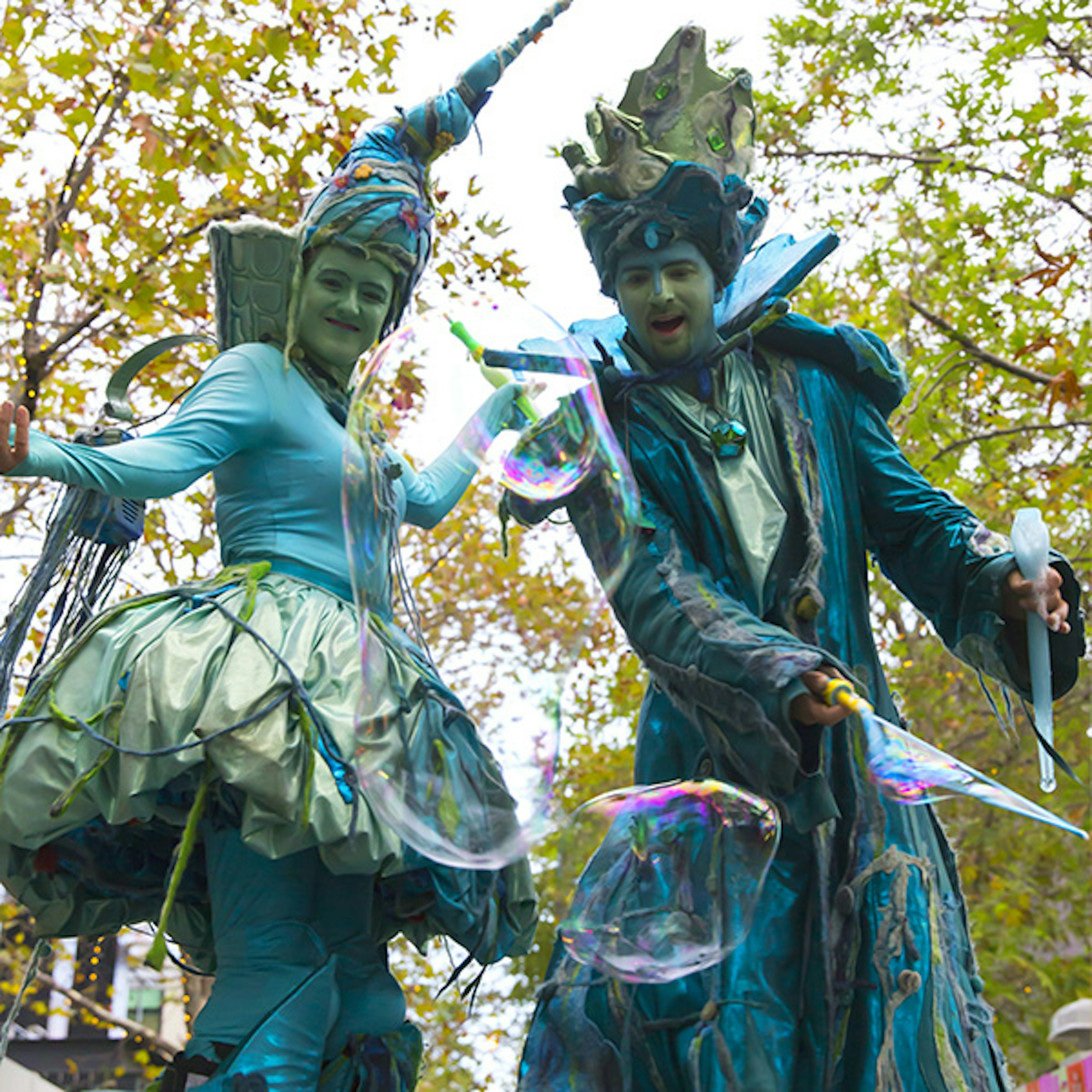 Two Water Drop Stilt Walkers are dressed in aqua coloured costumes playing with bubbles