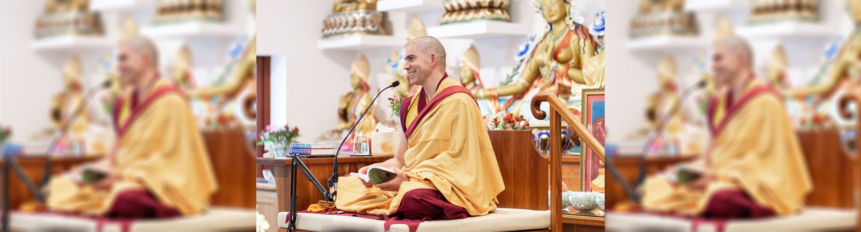 A Buddhist monk is sitting on stage wearing robes and smiling while talking into a microphone