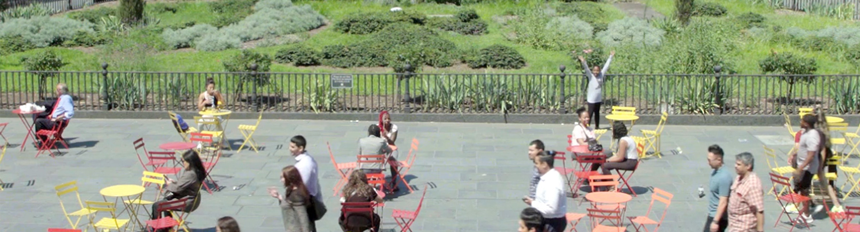 People are sitting on small colourful tables and chairs outside in a public space with a green park in the background