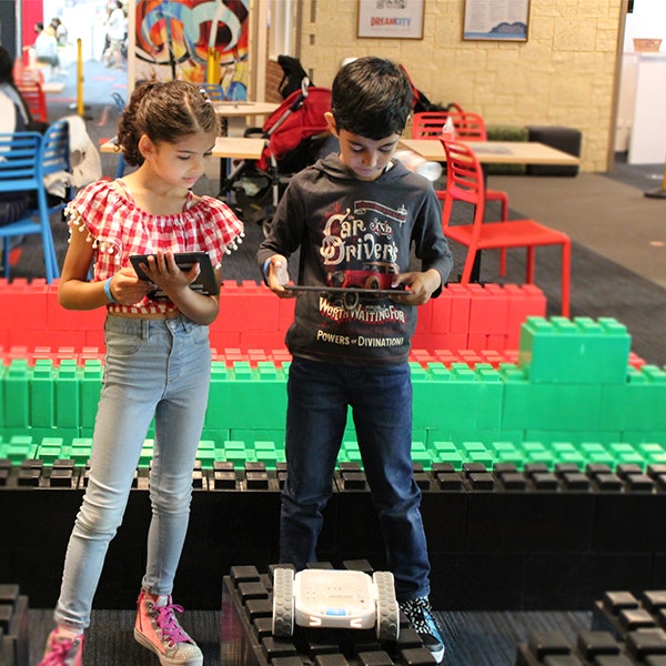Two kids looking at iPads while controlling a robot in a large Lego maze
