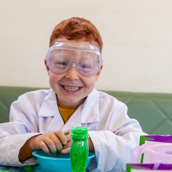 A child in a lab coat and wearing protective goggles smiles at the camera for the Silly Scientist Fizz Kidz workshop
