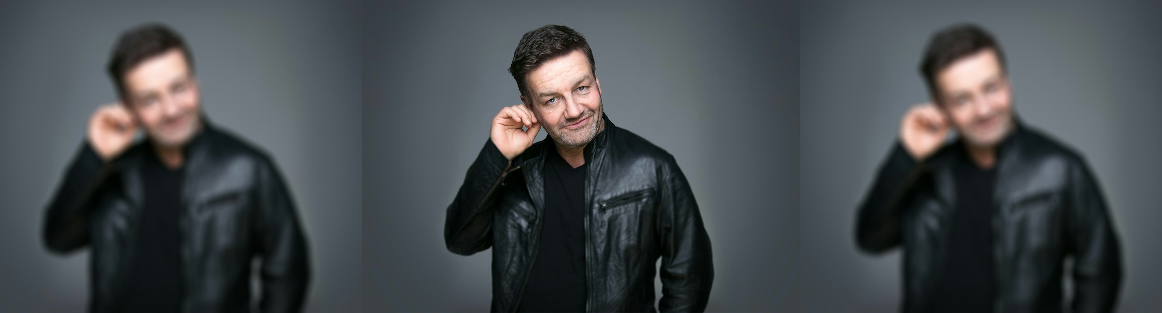 Lawrence Mooney smiling at the camera wearing a black leather jacket while he is rubbing his right ear