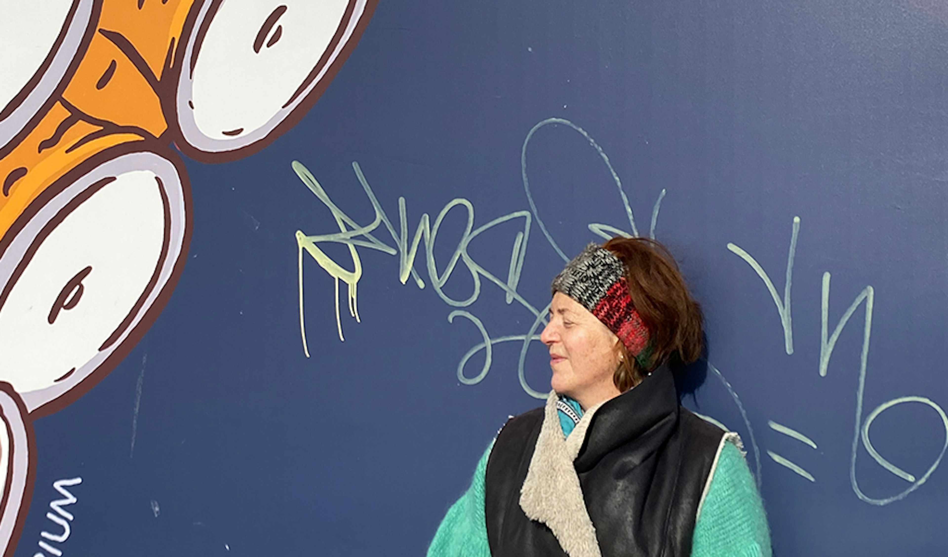 Anita is standing with her back to a navy blue wall with some graphic tags scrawled in yellow, her face is slightly upward and her eyes are closed due to the sunshine, in the top left corner is a part of a large octopus tentacle painted on the wall