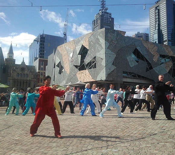 A group of people doing a Tai Chi pose in Fed Square while all wearing different coloured loose satin clothing