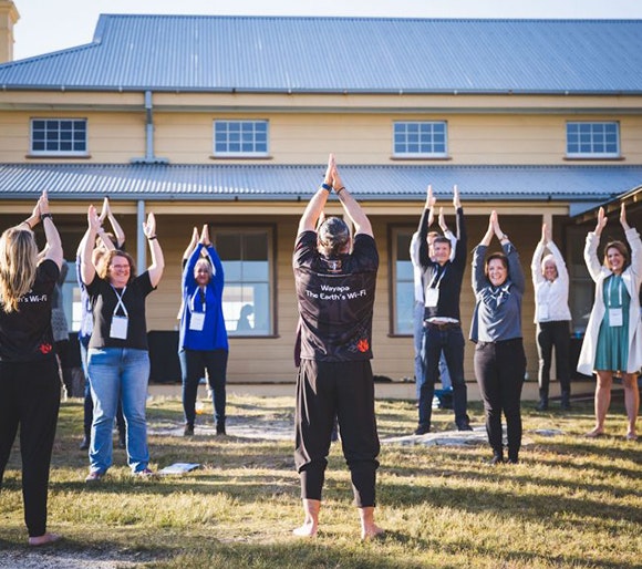 A group a people in front of a house in a yoga pose with their hands above their head with their palms together facing each other
