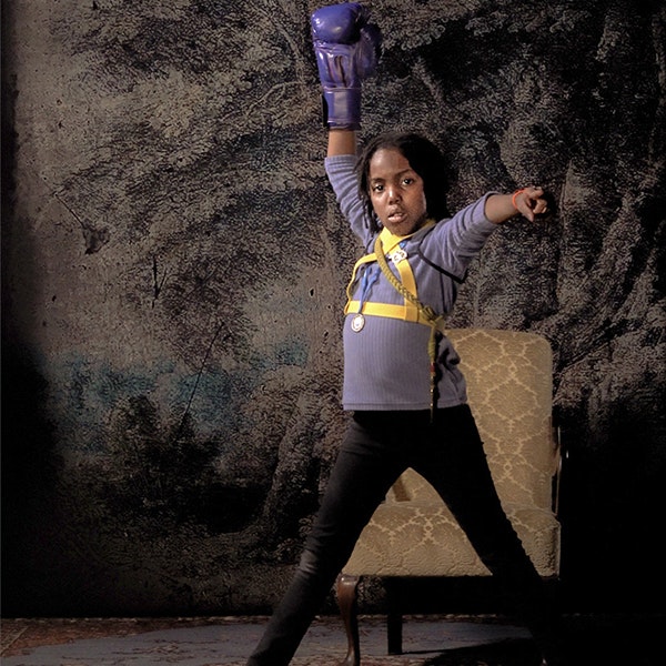A young girl has her right hand directly up in the air wearing a purple boxing glove, and her other hand is pointing towards the camera. She is wearing a purple shirt and black pants
