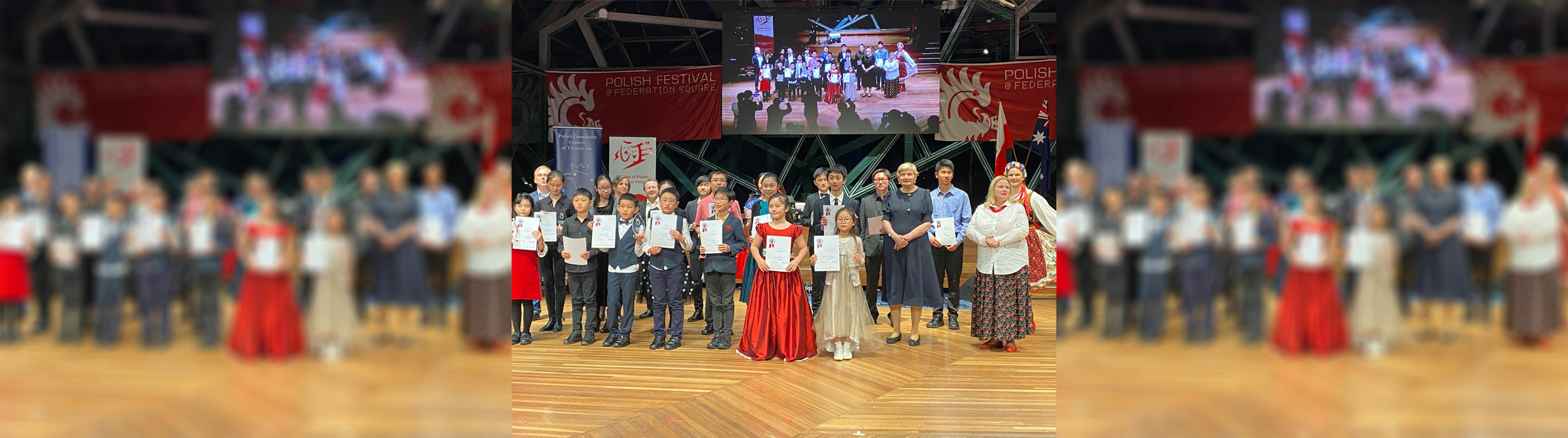 A group a children and adults on stage in the Edge dressed in formal attire posing for a photo after receiving awards for Polish Music Day