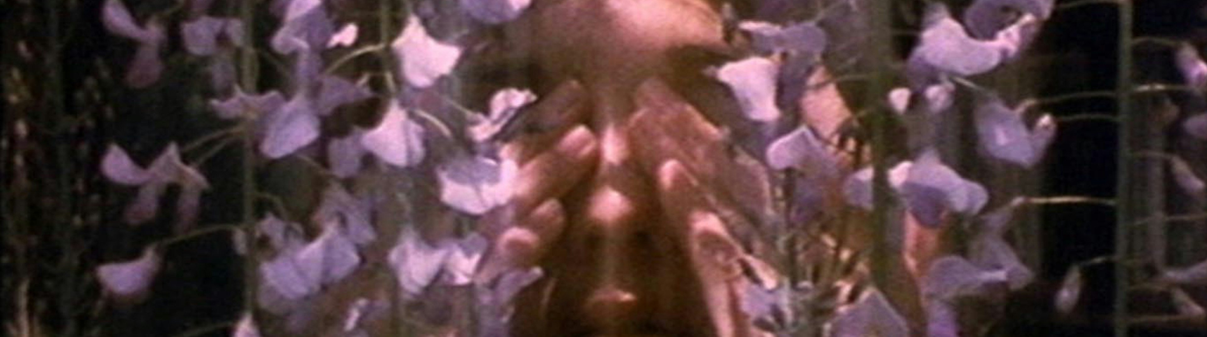 A grainy close up photo of a woman covering her eyes, with purple flowers all around her head