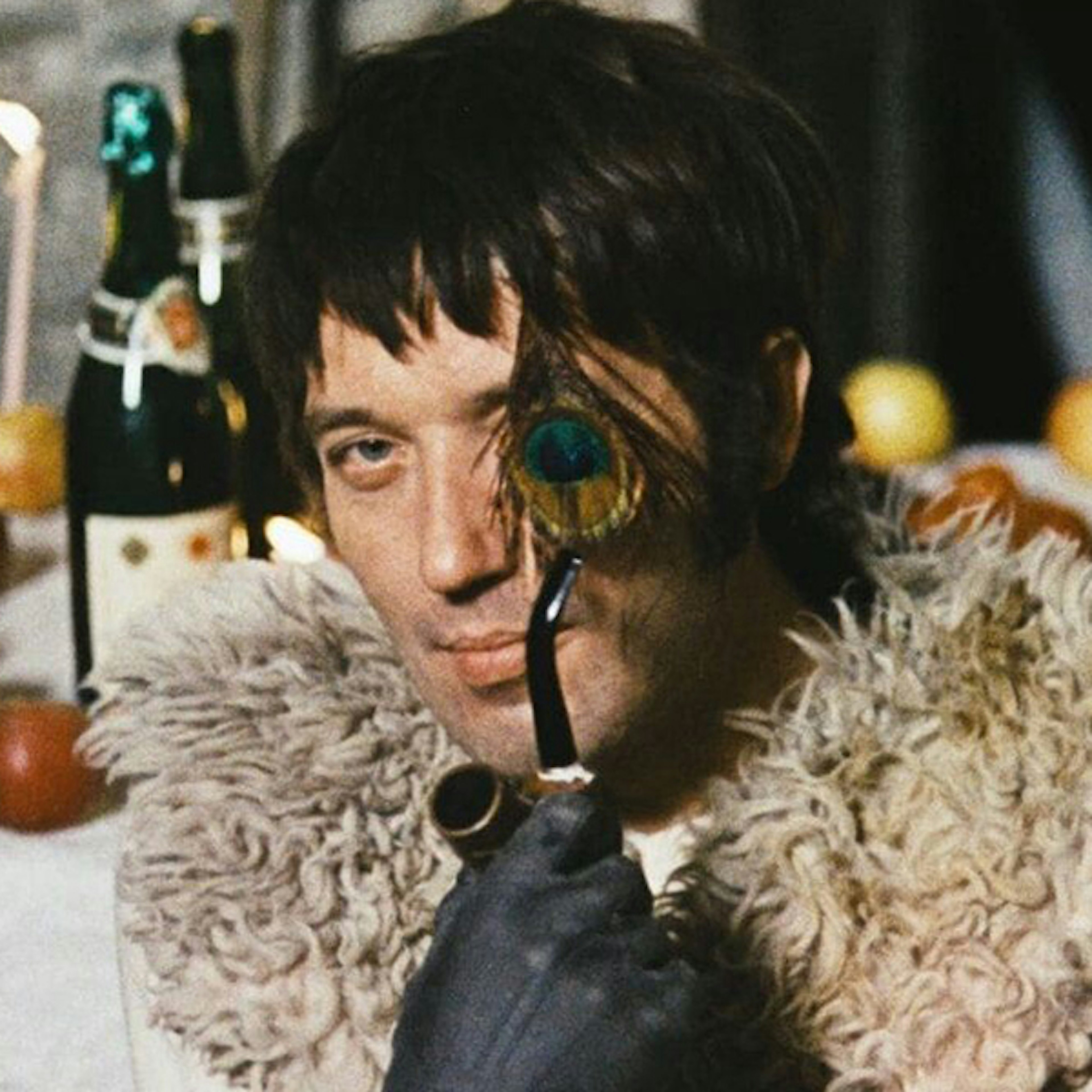 An image of a man with black hair looking at the camera holding a smoking pipe in a gloved hand, with a peacock feather covering his left eye, and a fur around his shoulders