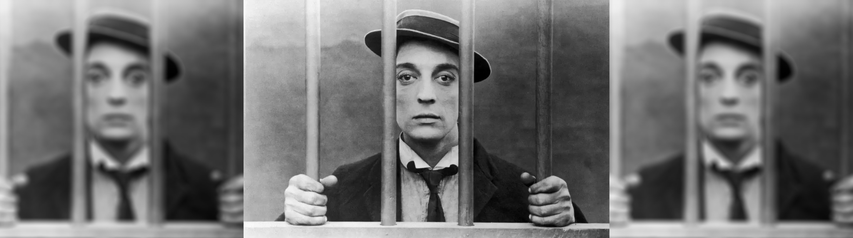 A film still from the black and white silent film, Cops, a man is wearing a flat brimmed 20s style hat, he has a sullen look and his behind bars