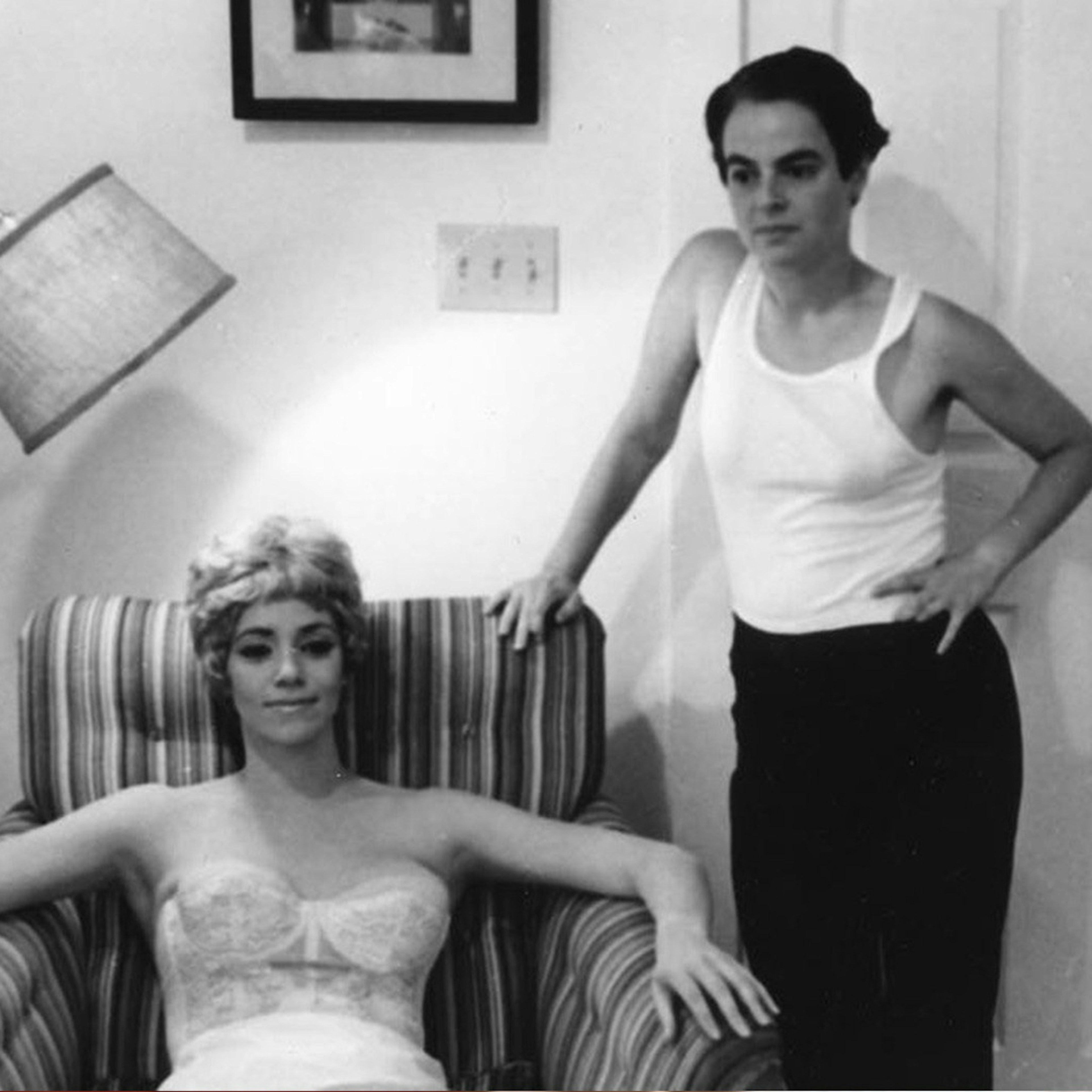 A black and white image of a woman on the right wearing a singlet standing next to an armchair with her left hand of her hip, there is another woman sitting on the striped armchair she is wearing a corset