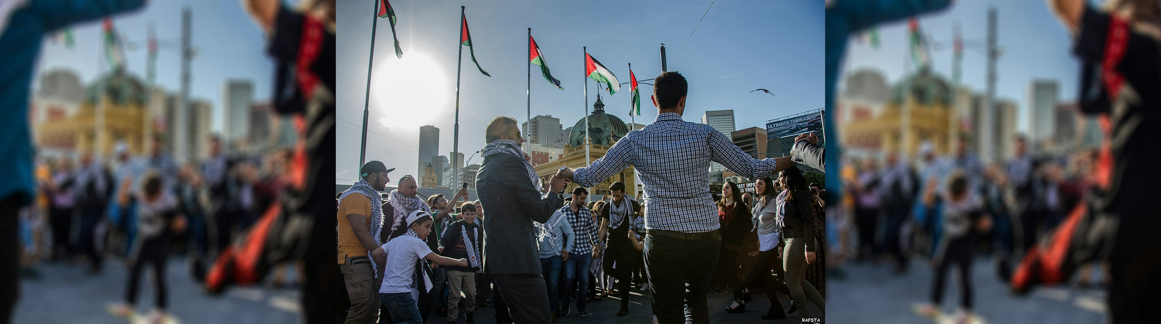 A group of Palestinian people holding hands dancing in a circle with five Palestine flags flying on poles in the air, and the top of Flinders Street Station in the background