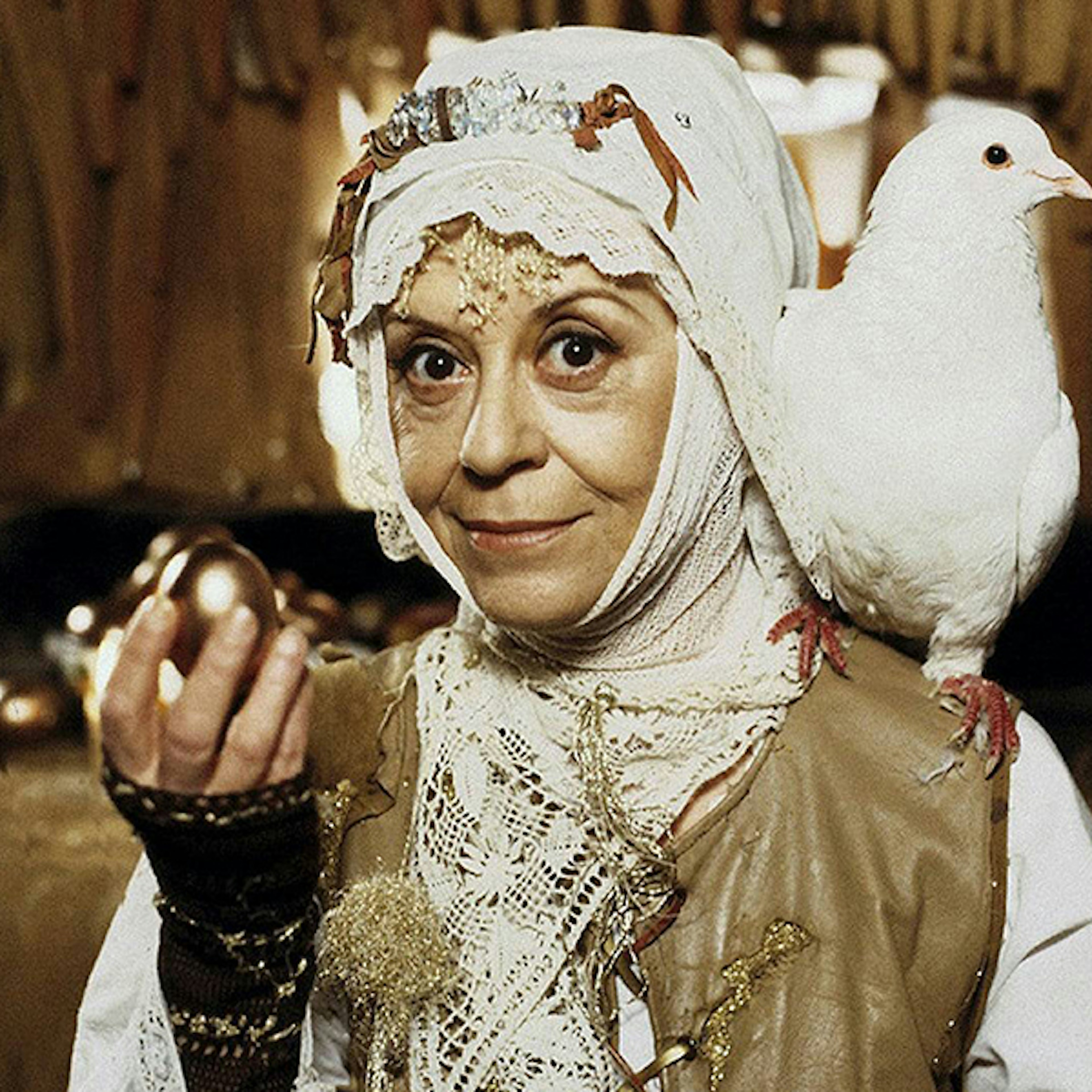 A woman looking whimsically into the camera holding a golden egg in her right hand and a white dove is sitting on her left shoulder, her head is covered in a tight white shawl