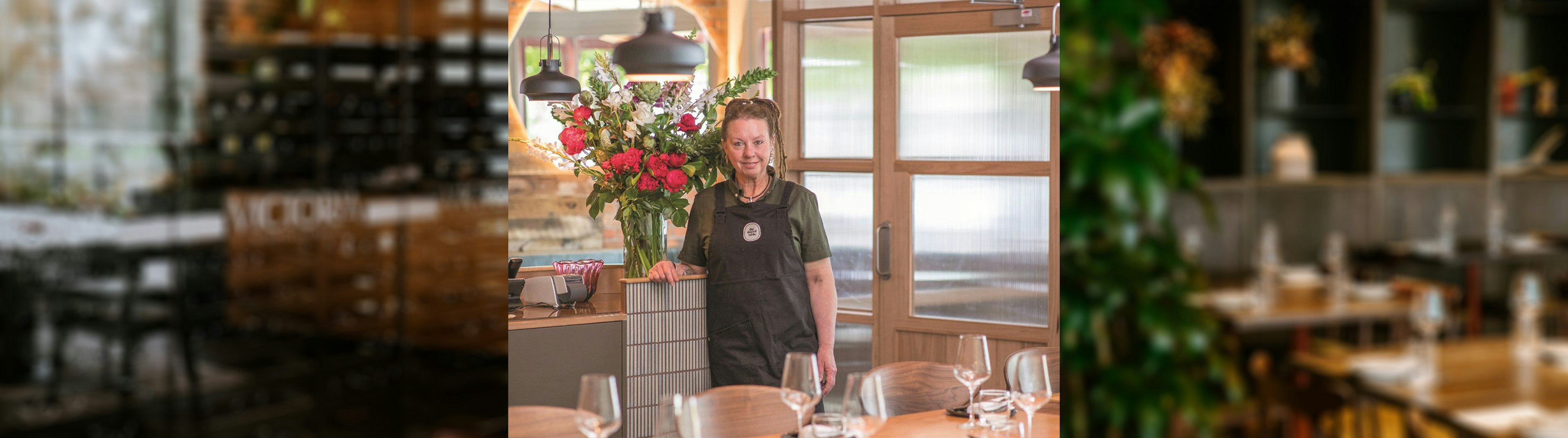 A woman with dreads tied up, standing in a restaurant next to a bench with a bunch of flowers to her right. She is smiling at the camera while wearing a dark grey apron over an olive green shirt