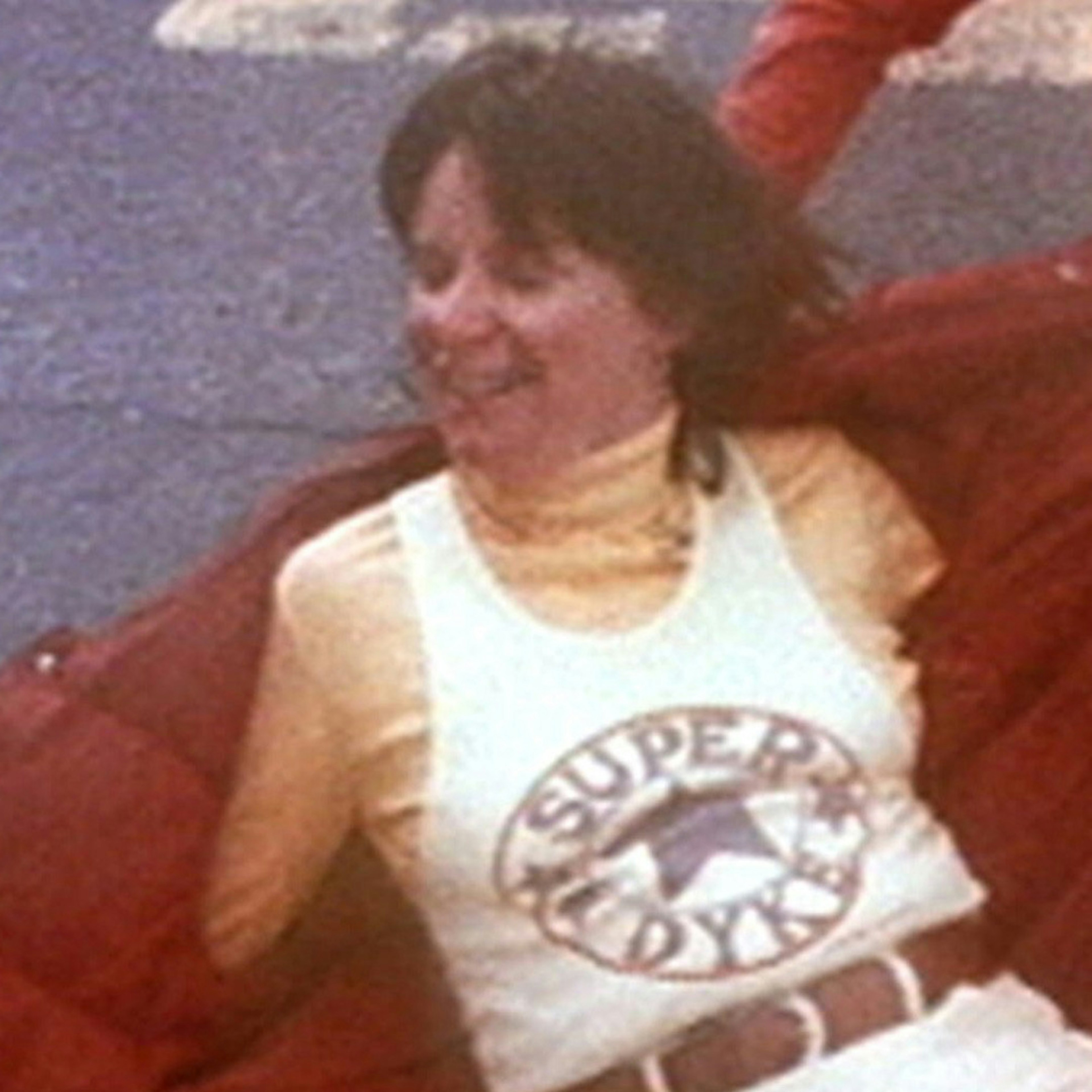 A photo of a woman in the 80s wearing a white singlet that says "Super Dyke" surrounding a blue star, underneath she is wearing a yellow skivvy and has a red puffer jacket open